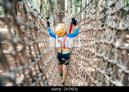 Boy walks across a mesh bridge, holding onto a cable. Back view Stock Photo