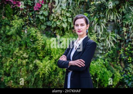 Self assured young female employee with dark hair in formal clothes standing in park near lush green bushes with crossed arms and looking at camera Stock Photo