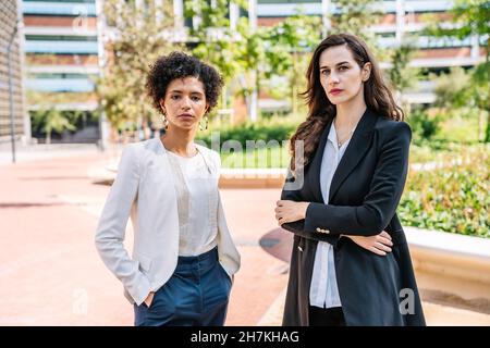 Serious confident young diverse female coworkers in elegant suits standing in city park with folded arms and hands in pockets and looking at camera Stock Photo