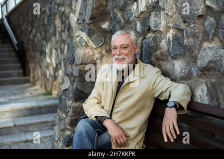 Smiling mature man sitting on bench by stone wall Stock Photo