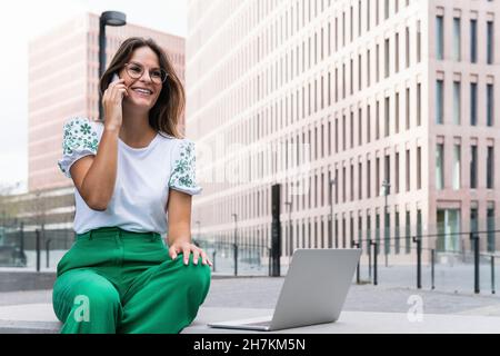 Smiling female professional with laptop talking on smart phone Stock Photo