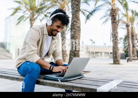 Smiling young man wearing wireless headphones using laptop on bench Stock Photo