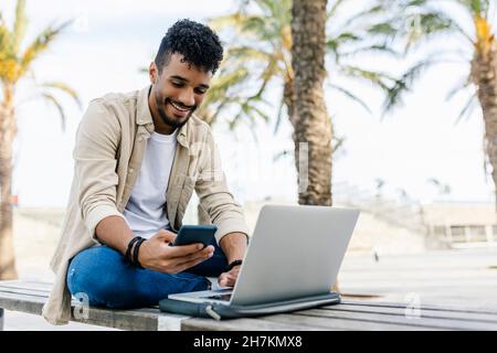 Smiling man with laptop using mobile phone on bench Stock Photo
