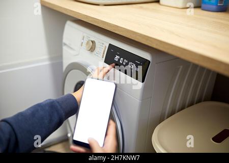Woman operating washing machine through smart phone in utility room at home Stock Photo