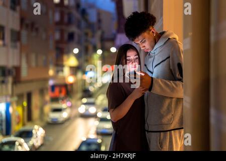 Multi-ethnic couple using smart phone while standing on balcony at night Stock Photo