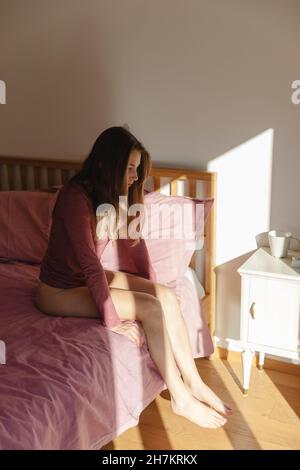 Sad young woman sitting alone on bed in bedroom Stock Photo