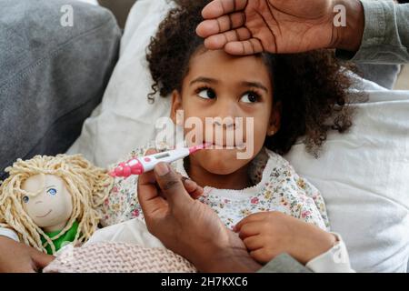 Father with thermometer measuring temperature of daughter on sofa Stock Photo