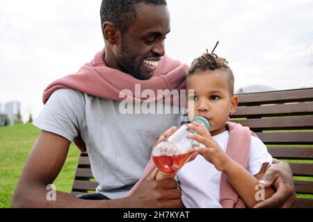 Smiling father looking at boy drinking juice on bench Stock Photo