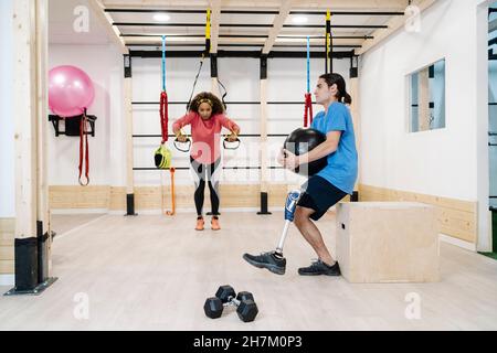 Smiling female instructor assisting disabled man doing push-ups on balance ball in gym Stock Photo