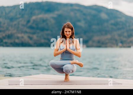 Smiling woman practicing toe stand on jetty in front of lake Stock Photo