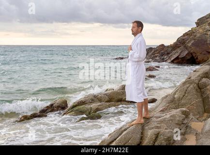 Man in bathrobe standing on rock at beach Stock Photo