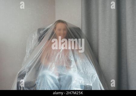 Playful woman covered in plastic sitting on armchair at new home Stock Photo