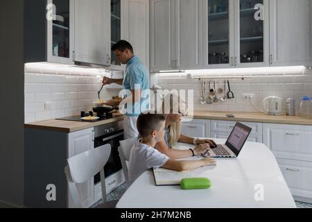 Mother helping son with homework on laptop at table by man cooking in kitchen Stock Photo