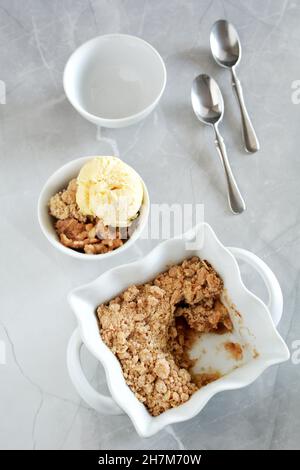 Fresh baked apple crumble in small casserole dish on light marble background in vertical orientation with bowl and ice cream. Stock Photo