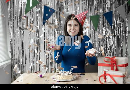 woman in a festive cap sits at the table at home and celebrates her birthday, confetti falls on her. woman portrait look into the camera. Stock Photo