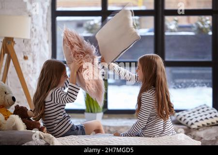 The image of two little sisters sitting on the bed in the room. They using pillow to fight each other Stock Photo