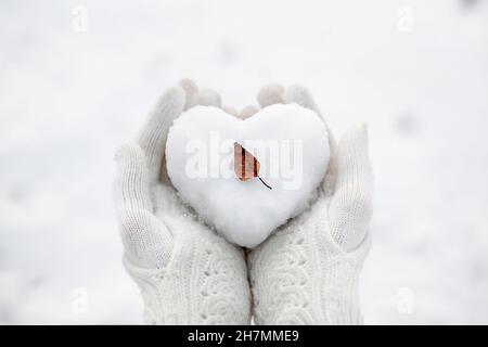 A snow heart with a small autumn leaf in the hands wearing knitted gloves against the background of a snow cover. Stock Photo