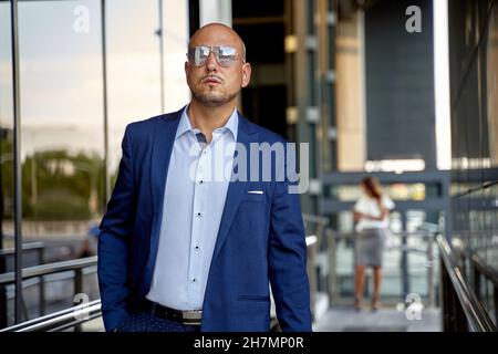 Bald businessman in blue jacket and stylish sunglasses holding hand in pocket and walking outside modern building while commuting to work Stock Photo