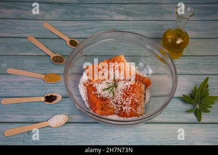 Salting salmon at home,  fillet in a transparent bowl olive oil and various spices in wooden spoons on the table Stock Photo