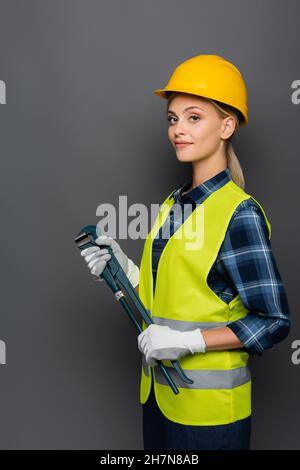 Young builder in helmet and safety vest holding pipe wrench isolated on grey Stock Photo