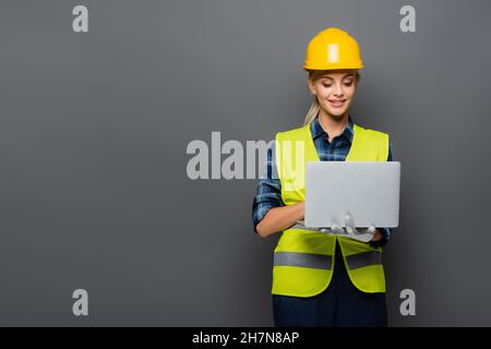 Positive builder in hardhat and protective vest using laptop isolated on grey Stock Photo