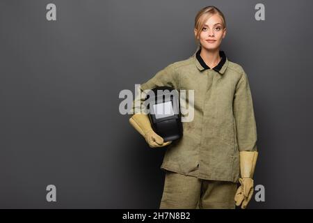 Blonde welder in gloves holding helmet on grey background Stock Photo