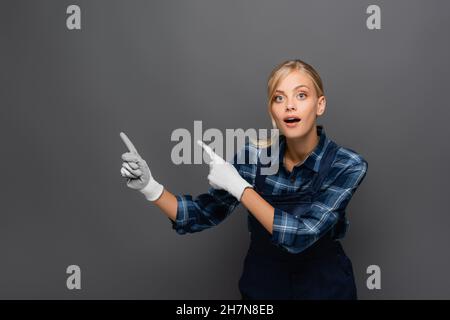 Astonished workwoman in gloves pointing with fingers isolated on grey Stock Photo