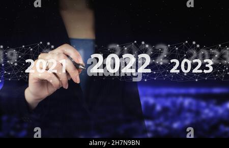 New year concept. 2022 new year. Hand holding digital graphic pen and drawing digital hologram 2022 sign on city dark blurred background. Happy New Ye Stock Photo