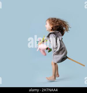 Toddler girl in grey dress jumps with hand made fabric stick horse on blue background Stock Photo