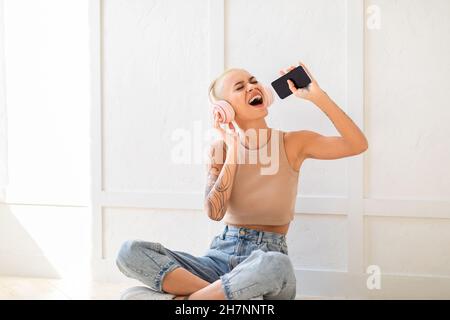 Joyful woman wearing wireless headphones and using cellphone as mic, singing while sitting on floor over white wall Stock Photo