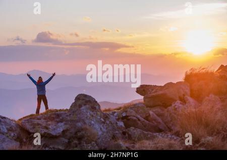 Lone tourist enjoying an amazing sunset in the mountains with raised arms. Focus on hiker figure Stock Photo