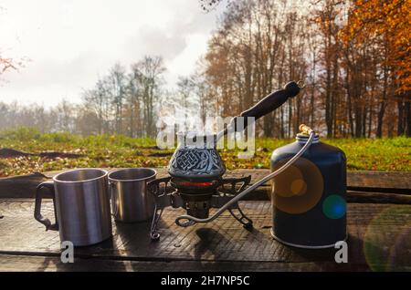 Boiling coffee cezve on the portable burner and metal mugs on th wooden table. Making hot drink outdoors in the park. Stock Photo