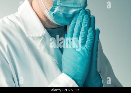 Medical professional wearing protective gloves and face mask with hands in praying and pleading position, selective focus Stock Photo