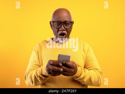 Emotional senior African American man in glasses looking at his smartphone in shock on orange studio background Stock Photo