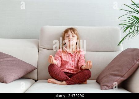 Little caucasian 3 year girl meditates on sofa while practicing yoga. Calm Kid Girl Daughter in pajamas with close eyes meditates in morning in home Stock Photo