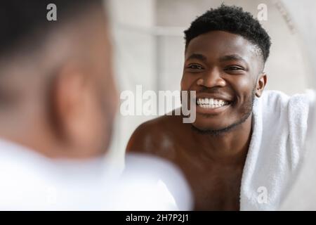 African Guy Looking At Toothy Smile In Mirror In Bathroom Stock Photo