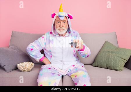 Photo of angry upset man pensioner unicorn sleepwear sitting sofa choosing tv program eating popcorn isolated pink color background Stock Photo