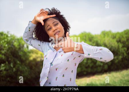 A young woman doing frame gesture with her hands to the camera and looking at camera with a background of green bushes Stock Photo