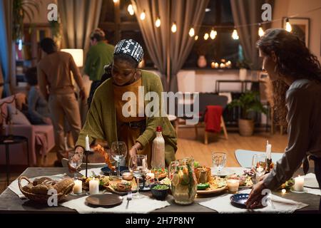 Two women setting the dining table for festive dinner with their friends in the living room Stock Photo