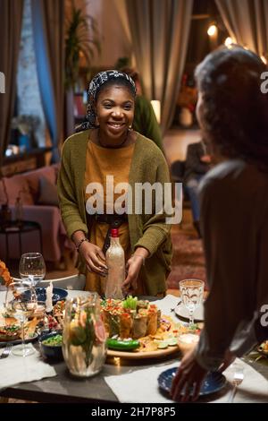 Happy African woman bringing bottle of wine for dinner and talking to friend while she setting the table Stock Photo