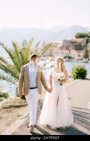 The groom and the smiling bride with a bouquet walk holding hands on the pier near the palm tree Stock Photo