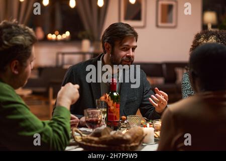 Young man sitting at dining table and talking to the people during dinner party at home Stock Photo