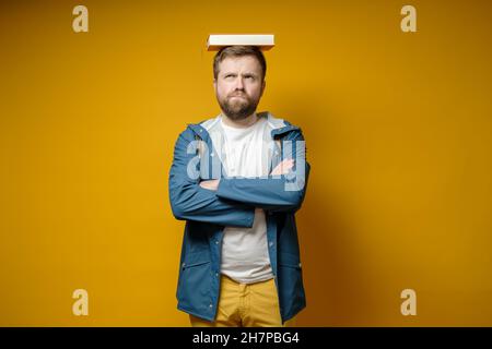 Pensive, funny male student put a book on his head as if it were a graduate hat and dreams of graduation. Yellow background. Stock Photo