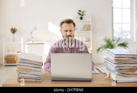Portrait of serious and focused manager working on laptop among piles of documents. Stock Photo