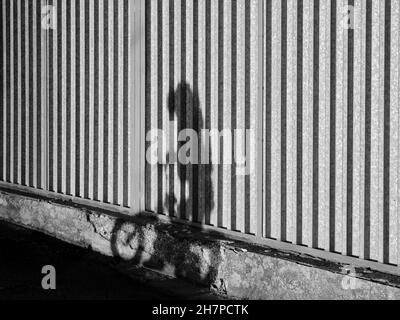 Black and white street photo of a child's shadow riding a bicycle on a striped metal wall. Stock Photo