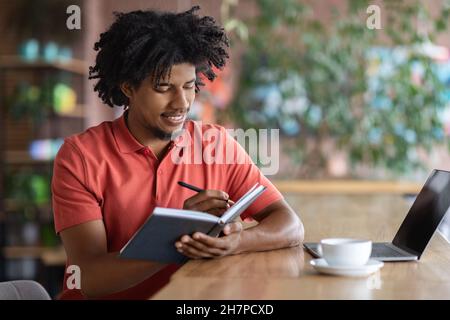 Time Management. Young Black Man Writing In Notepad While Sitting In Cafe Stock Photo