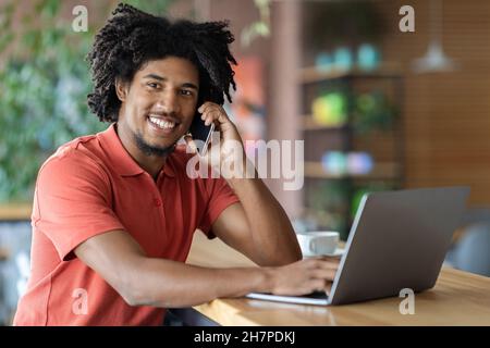 Modern Entrepreneur. Black Businessman Talking On Cellphone And Using Laptop In Cafe Stock Photo