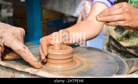 The hands of an old teacher and a young student in clay. The potter's wheel rotates the clay under the palms. Handmade, working with natural materials Stock Photo