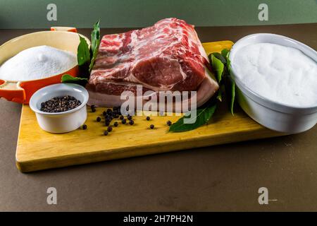 Pork loin or back joint with ingredients for dry curing into bacon, salt, sugar, peppercorns, juniper berries and bay leaves, landscape, on wooden cho Stock Photo