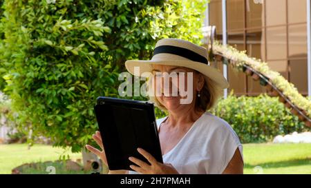 keen Senior female blonde businesswoman in hat uses tablet pc, digital tablet for business work or study in her own green garden. woman aged 50-55 Stock Photo